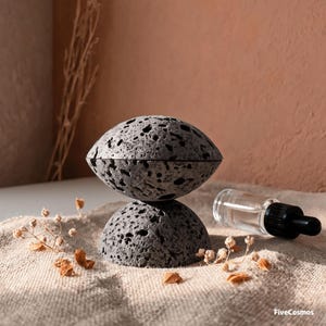 以下が含まれることがあります： 2つの積み重ねられた丸みを帯びた質感のある石のパーツで構成された、ダークグレーの多孔質な火山岩ディフューザー。黒いスポイトキャップが付いた小さな透明なガラス瓶が、石のオブジェクトの横に置かれています。
