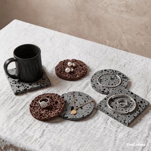 Puede incluir: Una taza negra, dos posavasos cuadrados grises moteados, dos posavasos redondos marrones porosos y dos posavasos redondos grises moteados. Las joyas incluyen pendientes de perlas, una pulsera de cadena, un collar dorado y dos anillos de plata.