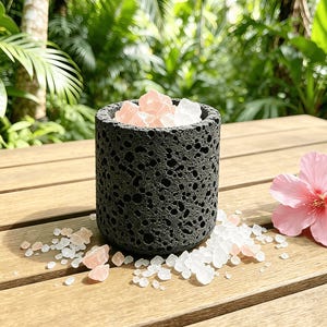 May include: A cylindrical, dark gray, porous stone container filled with pink and white crystals. The container sits on a wooden surface, with scattered crystals and a pink flower nearby. The background features lush green foliage.