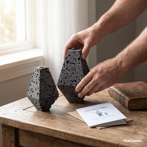 May include: Two geometric, dark gray, porous volcanic rock vessels with a textured, speckled surface. A white greeting card with the text FATHER'S DAY and a small illustration of two figures is placed next to the vessels on a wooden surface.