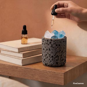 May include: A cylindrical dark gray porous stone diffuser filled with translucent blue and clear crystals. A small glass dropper bottle with a black cap sits on a stack of three books, while a hand holds a second dropper dispensing liquid.
