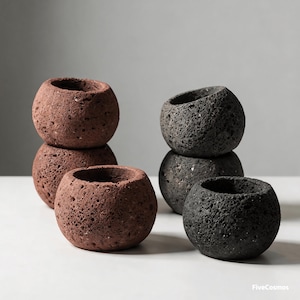 May include: A collection of five spherical, porous planters with a rough, volcanic texture. Three planters are reddish-brown and two are dark charcoal gray. The planters are arranged in two stacks with one in front. The text FiveCosmos is visible.