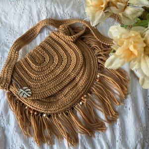 May include: A handmade, crochet shoulder bag in a warm tan color. The bag features a semi-circular design with a long strap and fringe detailing along the bottom. A silver leaf charm hangs from the front, and wooden beads adorn the fringe.