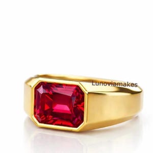Peut inclure: Une bague en or ornée d'une pierre précieuse rouge foncé de forme rectangulaire. La bague a un design simple et moderne avec une finition polie. Le texte "Lunoviamakes" est visible sur le côté de la bague.