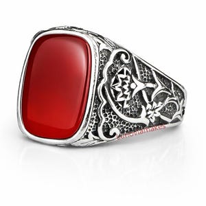 Peut inclure: Une bague en argent avec une pierre rouge vif de forme rectangulaire. La bague présente des motifs complexes et détaillés le long de l'anneau, mettant en valeur un design classique. La pierre mesure environ 2 cm de long et 1,3 cm de large.
