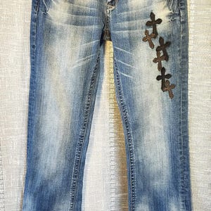 May include: Blue denim jeans with a faded wash, featuring a button and zipper fly closure, five pockets, and a series of dark brown cross-shaped patches embroidered vertically along the upper left thigh area. The waistband includes a visible brand label.