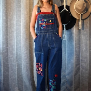 May include: Dark blue denim overalls featuring a floral fabric patch on the chest pocket, side pockets, and lower leg. The overalls include red lace trim along the chest, yellow buttons, and a floral embroidered stem with a red flower on the leg.