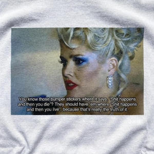 Könnte beinhalten: Weißes T-Shirt mit einer Grafik einer Frau mit auffälligem Make-up und einem Zitat über das Leben. Das Zitat lautet: „Du kennst doch diese Aufkleber, auf denen steht: 'Scheiße passiert und dann stirbst du'? Sie sollten 'Scheiße passiert und dann lebst du' haben…“