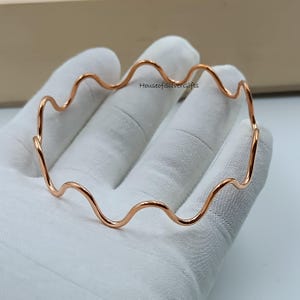 Può includere: Un braccialetto a bracciale ondulato color rame. Il braccialetto ha un design a onda continua. Il braccialetto è esposto su una superficie bianca. Il testo "HouseofSilverGifts" è visibile sulla superficie.