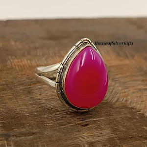 May include: A silver ring featuring a large, teardrop-shaped, fuchsia-colored gemstone. The gemstone is set in a detailed silver bezel. The ring has a split shank design. The text "HouseofSilverGifts" is visible on the ring.