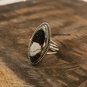 Può includere: Un anello d'argento con una pietra di forma ovale. La pietra presenta un mix di colori bianco, marrone e verde acqua. L'anello ha un design a corda intrecciata attorno alla pietra e una base ad anello a tre bande. Il testo "HouseofSilverGifts" è visibile.