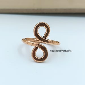 May include: A rose gold-toned ring featuring an open, bypass-style design with two circular loops at the ends. The metal surface is smooth and reflective. The text Houseofsilver&gifts is printed in black near the right side of the ring.