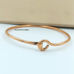 Puede incluir: Una pulsera rígida de metal en tono oro rosa con un mecanismo de cierre tipo gancho. La superficie lisa y pulida de la pulsera refleja la luz, y el texto Houseofsilver&gifts es visible cerca de la parte superior de la banda circular.