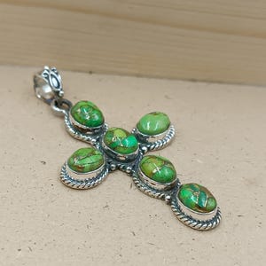 May include: A silver-toned cross pendant with five oval-shaped, green turquoise stones. The stones have a matrix of gold and brown. The pendant has a decorative bail and a detailed, rope-like border around each stone.