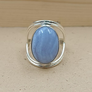 Puede incluir: Un anillo de plata con una gran piedra preciosa de calcedonia azul claro de forma ovalada con sutiles bandas blancas. La banda consta de dos hilos de plata paralelos que se curvan alrededor de la piedra central. El texto HouseofSilverGifts es visible.