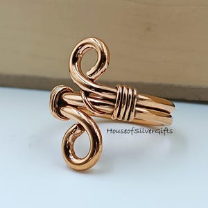 May include: A copper-colored wire ring with a unique design. The ring features two swirling loops and a wrapped wire detail. The band is adjustable. The text "HouseofSilverGifts" is visible on the ring.