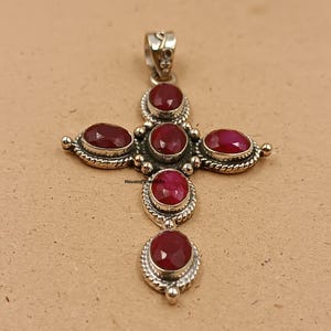 Puede incluir: Colgante de cruz de plata con seis piedras preciosas de color rojo intenso, ovaladas y redondas. El colgante tiene un diseño detallado y ornamentado con un lazo en la parte superior para una cadena. Las piedras preciosas están engastadas en un bisel de plata.