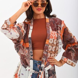 Peut inclure: Une femme porte un kimono à motifs avec des motifs patchwork marron, orange et blanc. Le kimono est ceinturé à la taille. Elle porte des lunettes de soleil avec une monture marron clair et des verres foncés. Le fond est blanc uni.