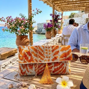Peut inclure: Une pochette orange et grise à motifs avec une chaîne dorée et une pampille est posée sur une table en bois. Des coquillages et une fleur blanche sont éparpillés autour du sac. L'arrière-plan montre la mer et des personnes.