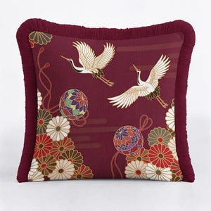 Könnte beinhalten: Burgunderfarbenes quadratisches Kissen mit dekorativem Fransenbesatz. Das Kissen zeigt ein japanisch inspiriertes Design mit weißen Kranichen im Flug, bunten Blumenmustern und gemusterten Kugeln.
