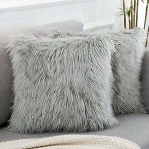 Könnte beinhalten: Zwei quadratische, flauschige, graue Zierkissen mit langer, zotteliger Kunstfellstruktur werden auf einem grauen Sofa gezeigt. Die Kissen sind nebeneinander positioniert und schaffen eine gemütliche und einladende Ästhetik. Die Kissen verleihen dem Wohnzimmer einen Hauch von Luxus.