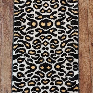May include: A rectangular rug with a repeating leopard print pattern. The rug features black spots with gold-coloured centres on a white background. The rug has a dark grey border and is placed on a wooden floor.