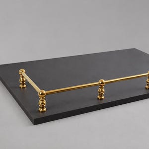 May include: A rectangular, dark gray tray with a gold-colored metal railing. The railing has a horizontal bar supported by decorative vertical posts with ball-shaped finials. The tray is a simple, elegant design.