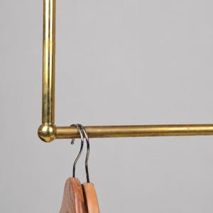 May include: A gold-coloured metal clothing rack with a rectangular frame. Two wooden hangers hang from the horizontal bar. The rack is designed for organising clothing. The background is a solid, neutral colour.