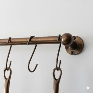 May include: A bronze-colored metal kitchen rail with several hanging hooks. The rail is mounted on a white wall and has a decorative end piece. The hooks are designed to hold kitchen utensils or other items.