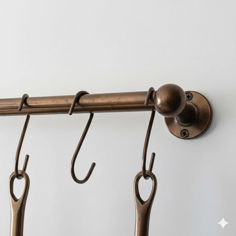 May include: A bronze-colored metal kitchen rail with several hanging hooks. The rail is mounted on a white wall and has a decorative end piece. The hooks are designed to hold kitchen utensils or other items.