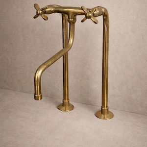 May include: A vintage-style brass faucet with a curved spout and two cross-shaped handles. The faucet has a warm, golden hue and is mounted on a gray surface. The design is reminiscent of classic bathroom fixtures.