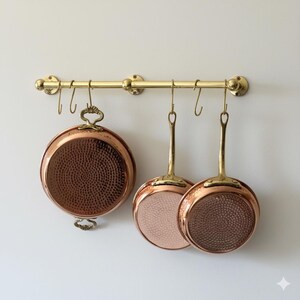 May include: A brass pot rack with three copper pans hanging from it. The pans have copper exteriors and interiors, with brass handles. The pans have a hammered texture. The pot rack is mounted on a white wall.