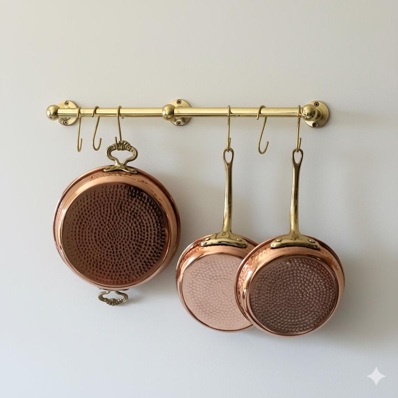 May include: A brass pot rack with three copper pans hanging from it. The pans have copper exteriors and interiors, with brass handles. The pans have a hammered texture. The pot rack is mounted on a white wall.