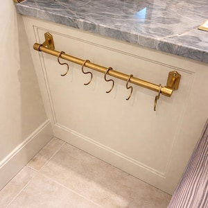 Op de afbeelding: Een goudkleurige metalen stang met zes haken, gemonteerd op een witte kast. De stang is met decoratieve beugels aan de kast bevestigd. Het aanrechtblad boven de kast is van grijs marmer.