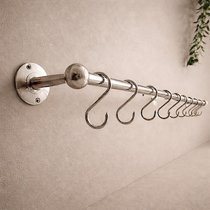 Könnte beinhalten: Eine verchromte Küchenreling mit S-förmigen Haken. Die Reling ist an der Wand befestigt und hat eine dekorative Kugel in der Mitte. Die Haken sind gleichmäßig verteilt und eignen sich zum Aufhängen von Küchenutensilien oder anderen Gegenständen. Ca. 30 cm lang.