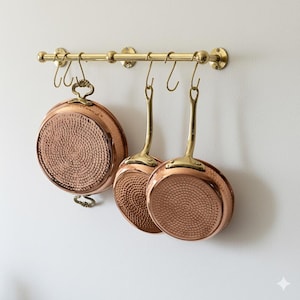 May include: A brass pot rack with three copper pans hanging from it. The pans have brass handles and a hammered texture on the bottom. The pot rack is mounted on a white wall.