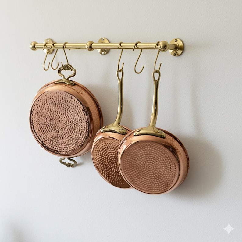 May include: A brass pot rack with three copper pans hanging from it. The pans have brass handles and a hammered texture on the bottom. The pot rack is mounted on a white wall.