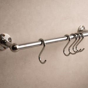 Op de afbeelding: Een verchroomde keukengerei-rek met vijf S-haken. Het rek is aan een muur bevestigd en heeft een horizontale stang met decoratieve uiteinden. De haken zijn ontworpen om keukengerei op te hangen.