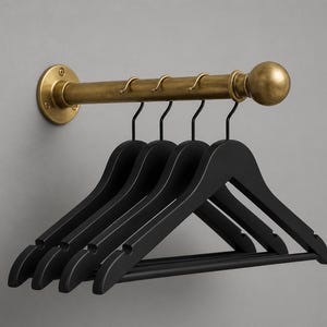 May include: A wall-mounted clothes rail in a brass finish, holding five black wooden coat hangers. The rail has a horizontal bar with hooks and a decorative ball on the end. The hangers are evenly spaced.