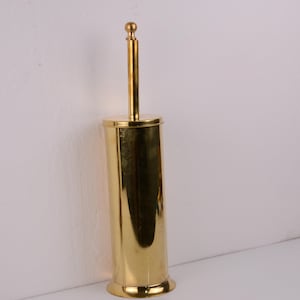 Puede incluir: Un cepillo y soporte para inodoro de color dorado. El soporte es cilíndrico con una base redondeada y una tapa. El mango del cepillo se extiende hacia arriba, rematado con un pequeño remate redondo. El artículo es de metal brillante.