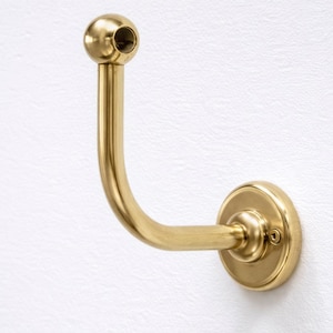 Op de afbeelding: Een messing wandhaak met een gebogen ontwerp. De haak heeft een ronde basis, een gebogen arm en een kogelvormig uiteinde met een gat. Het is gemonteerd op een witte, getextureerde muur.