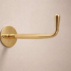 May include: A gold-colored, wall-mounted toilet paper holder. The holder has a circular base and a curved arm with a rounded end. The fixture is made of metal and is mounted on a light-colored wall.