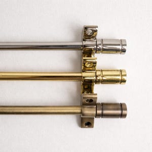 May include: Three curtain rods in different metallic finishes: silver, gold, and bronze. Each rod is mounted on a white wall with matching brackets. The rods are cylindrical and have decorative end caps.