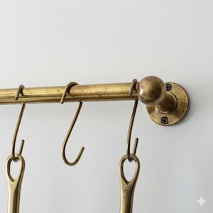 Pode incluir: Prateleira de utensílios de cozinha em latão envelhecido com ganchos para pendurar. A prateleira é montada em uma parede branca. Os ganchos são projetados para guardar utensílios de cozinha. O latão tem uma aparência vintage.