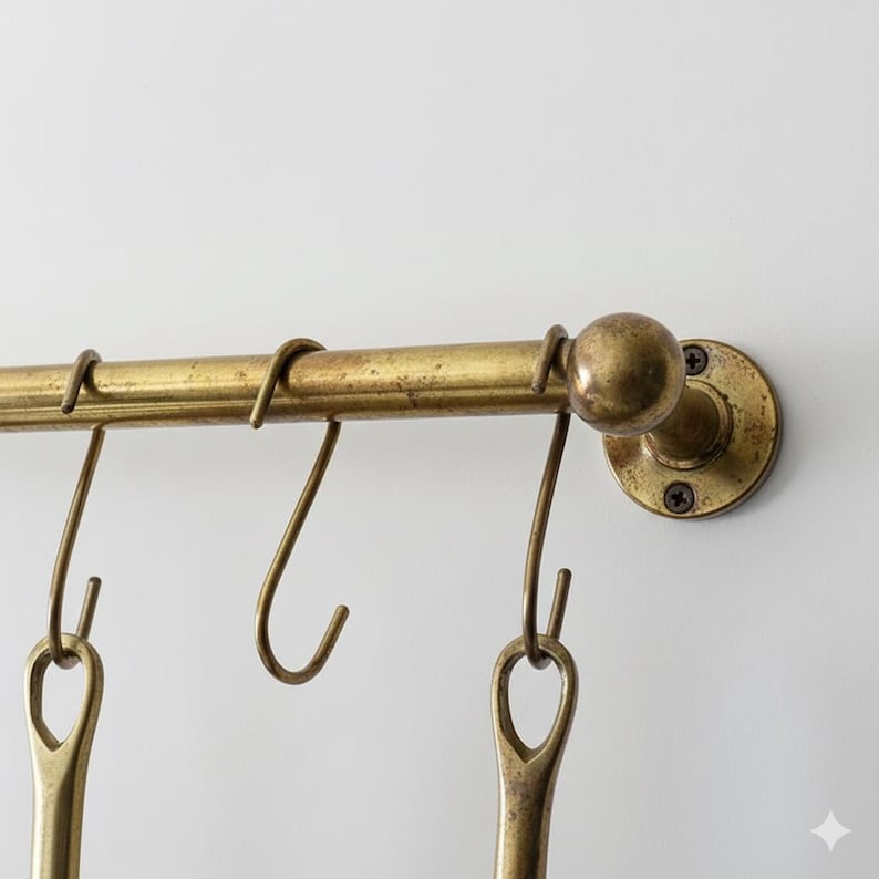 May include: Aged brass kitchen utensil rack with hanging hooks. The rack is mounted on a white wall. The hooks are designed to hold kitchen utensils. The brass has a vintage, antique appearance.