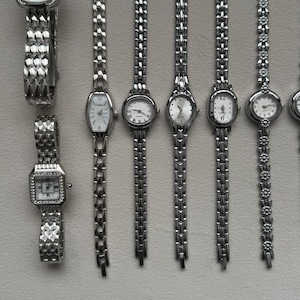 May include: A collection of silver-toned wristwatches with various face shapes, including rectangular, oval, and triangular. The watch bands are primarily linked metal, with some featuring decorative elements. The watch faces are white with black numerals and hands.