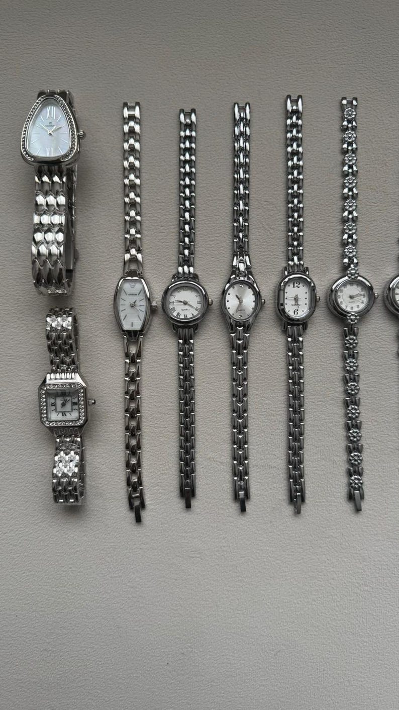 May include: A collection of silver-toned wristwatches with various face shapes, including rectangular, oval, and triangular. The watch bands are primarily linked metal, with some featuring decorative elements. The watch faces are white with black numerals and hands.