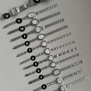 May include: An assortment of thirteen silver-toned wristwatches with various band designs and face shapes. The watches have white faces with black numerals and hands. The bands feature link and chain styles. The watches are displayed on a light gray surface.