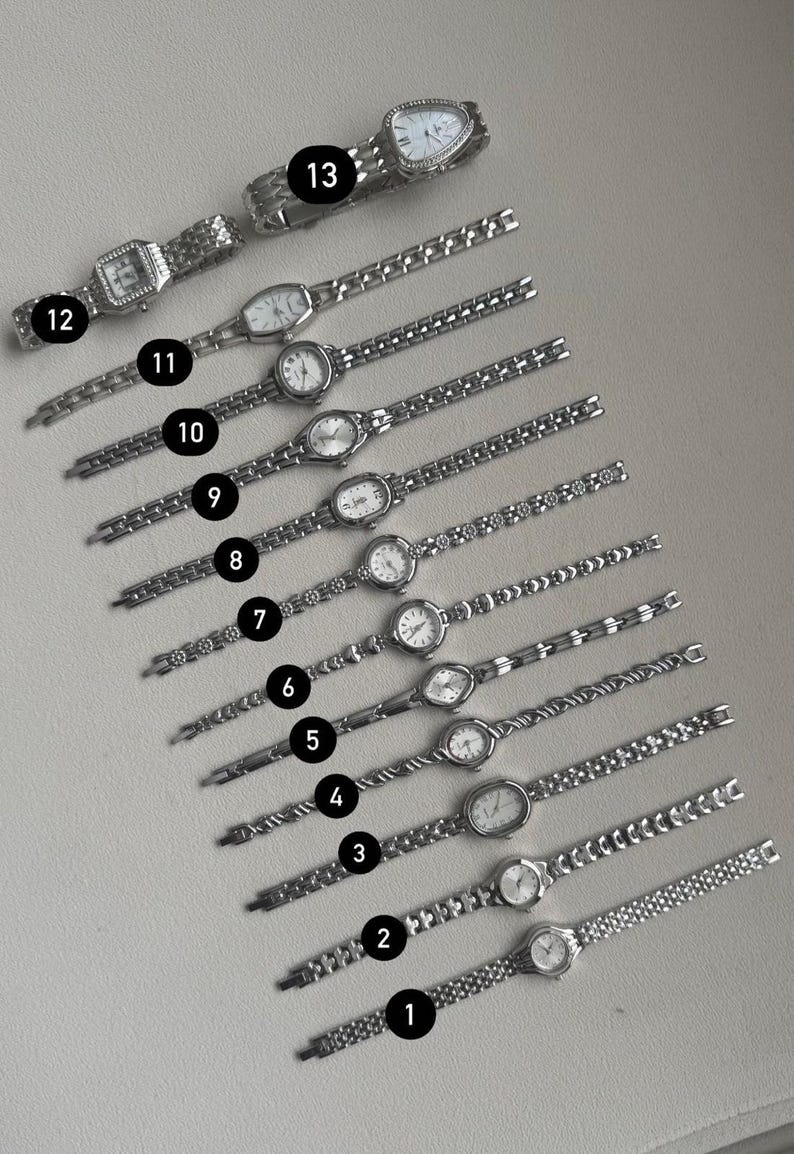 May include: An assortment of thirteen silver-toned wristwatches with various band designs and face shapes. The watches have white faces with black numerals and hands. The bands feature link and chain styles. The watches are displayed on a light gray surface.