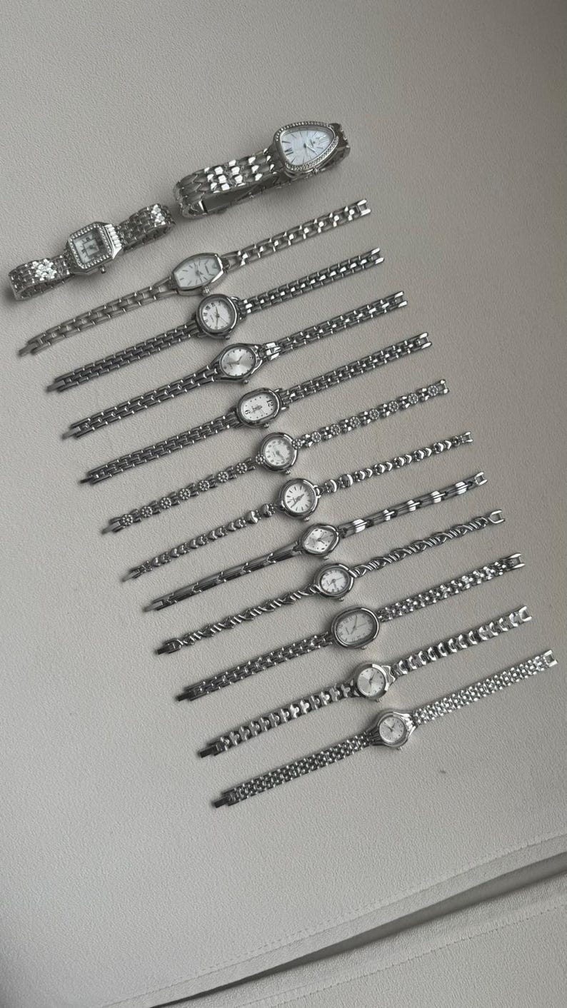 May include: An assortment of silver-toned wristwatches with various band designs and watch face shapes. The watches are arranged diagonally on a light-colored surface, showcasing their metallic sheen and intricate details. The watch faces are white.